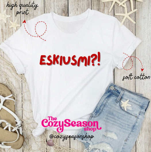 A white unisex T-shirt with the text 'ESKIUSMI?' printed in red, displayed flat with a pair of blue jeans and a pair of sandals on a wooden background. The shirt is presented with a graphic indicating the fabric is cotton blend and that it is machine washable.