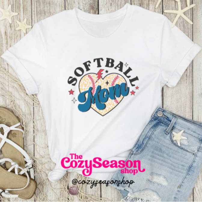 A white unisex T-shirt with 'SOFTBALL MOM' printed on the front, displayed flat with a pair of blue jeans and a pair of sandals on a wooden background, with decorative starfish and a small amount of sea foam.
