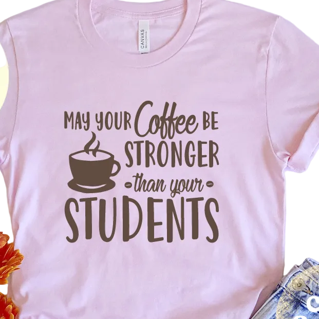 A pink unisex t-shirt with text graphic saying 'MAY YOUR COFFEE BE STRONGER THAN YOUR STUDENTS' on the front, displayed on a surface with decorative items around it.