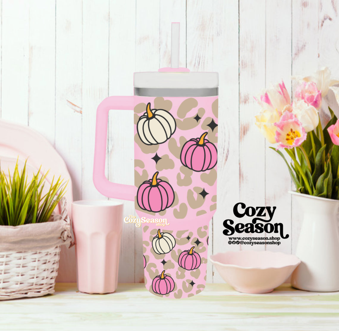 A 40oz pink stainless steel tumbler with a glitter print of pumpkins and leopard print, accompanied by a reusable straw and displayed on a wooden surface with a 'Cozy Season' label.