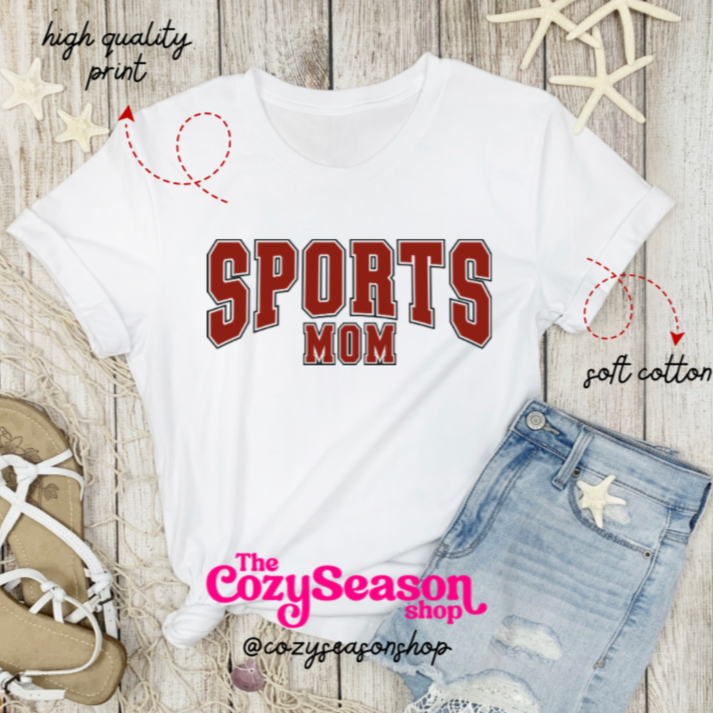 White t-shirt with 'SPORTS MOM' text on a wooden surface with denim jeans and sports equipment.
