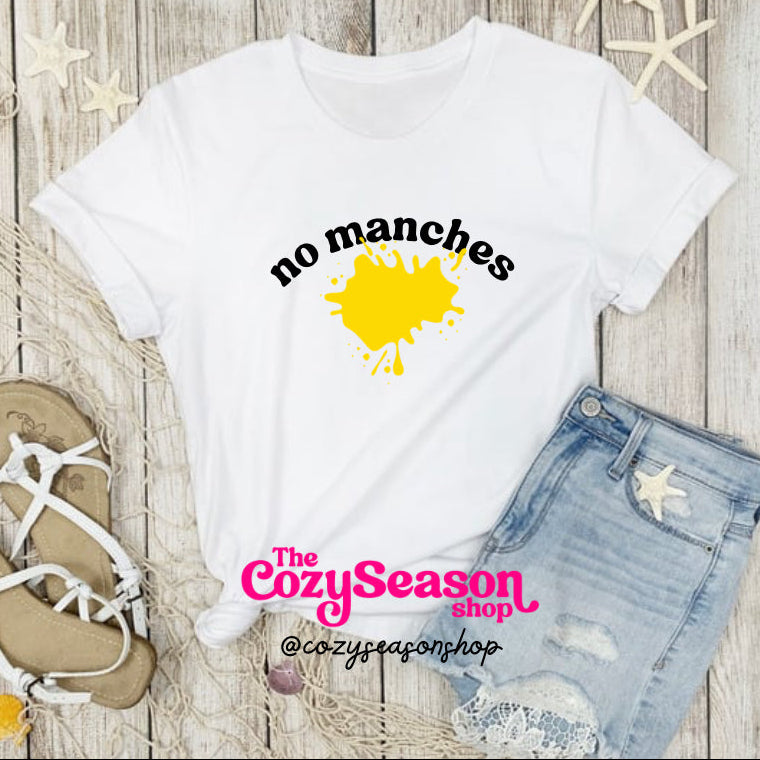 A white unisex T-shirt with the text 'NO MANCHES' printed in yellow, displayed on a wooden background with a pair of blue jeans, a pair of white sandals, and decorative starfish.