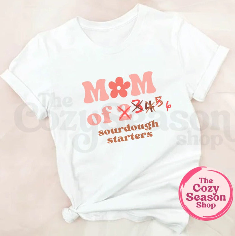 White t-shirt with pink text 'Mom of 2,3,4,5,6 sourdough starters' on a beige background, featuring 'The Cozy Season Shop' logo.