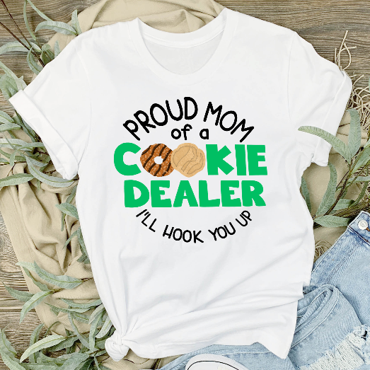 A white unisex t-shirt with text and a graphic design, displayed flat with a pair of blue denim jeans and a pair of white sneakers on either side, placed on a wooden surface with greenery in the background.