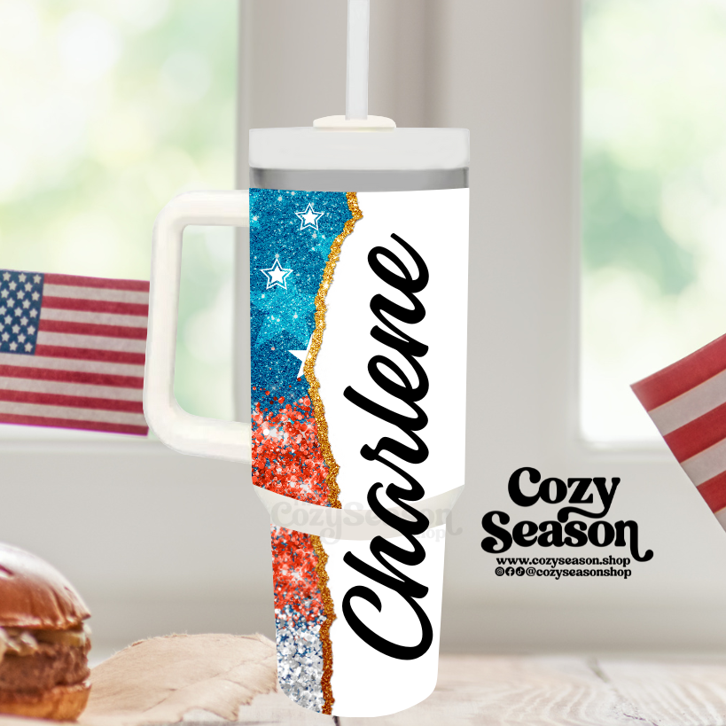 A 40oz iridescent stainless steel tumbler with a glitter print of the American flag and the name 'Chastine' on it, displayed on a table with a patriotic flag-themed design.