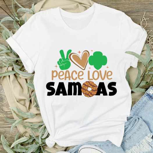 A white unisex adult t-shirt with the text 'PEACE LOVE SAMOAS' and a green leaf logo, displayed on a wooden background with a pair of blue jeans and a pair of white sneakers to the side.