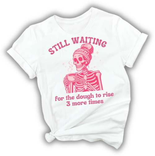 A white short-sleeve graphic T-shirt with a pink cartoon skeleton wearing a beanie and holding a mug, with the text 'STILL WAITING for the dough to rise 3 more times' printed on the front.