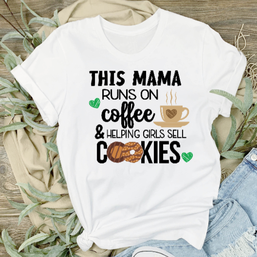 Adult unisex white t-shirt with text 'This mama runs on coffee & selling girl scout cookies' printed on it, displayed on a wooden surface with green foliage in the background, paired with blue jeans and white sneakers.
