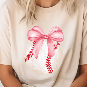 A woman wearing a beige t-shirt with a pink bow and red baseball print in the center.