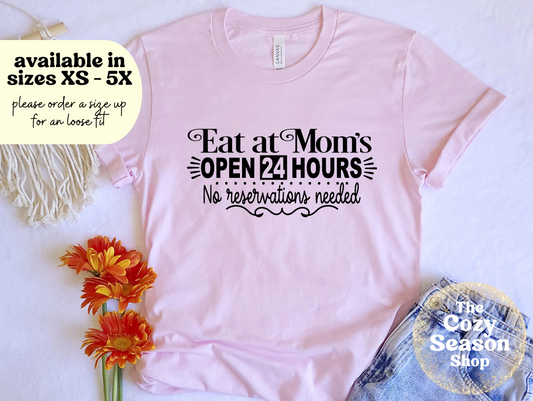 A pink unisex t-shirt with text 'EAT AT MOM'S OPEN 24 HOURS NO RESERVATIONS NEEDED' printed on it, displayed flat with a decorative flower and a fringed item to the side.