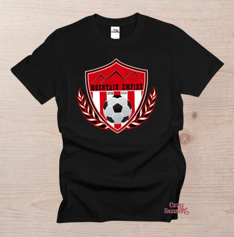 A black short-sleeve t-shirt laid flat on a light wood background, featuring a red and white shield graphic on the chest with mountains, a soccer ball, laurel leaves, and the text 'MOUNTAIN EMPIRE AYSO 930"