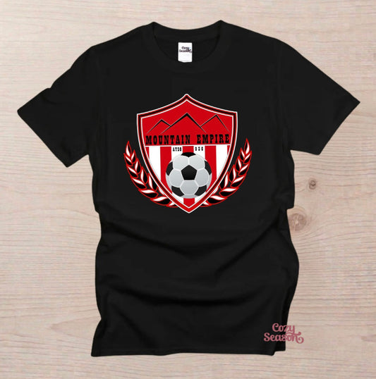 A black short-sleeve t-shirt laid flat on a light wood background, featuring a red and white shield graphic on the chest with mountains, a soccer ball, laurel leaves, and the text 'MOUNTAIN EMPIRE AYSO 930"