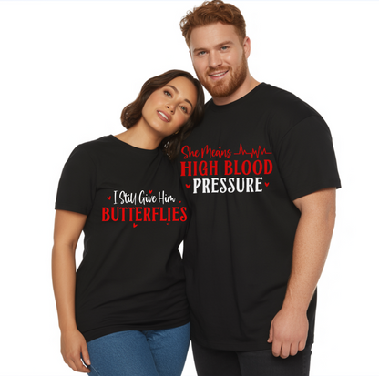 A COZY SEASON SHOP BRAND PHOTO OF A smiling couple wearing matching black t-shirts. The woman's shirt reads "I Still Give Him BUTTERFLIES" in white and red text with small hearts. The man's shirt reads "She Means HIGH BLOOD PRESSURE" with a red heartbeat graphic and white text.