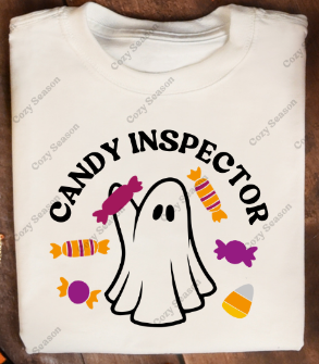 White crew neck t-shirt with 'Candy Inspector' text and a cartoon ghost graphic, displayed on a wooden surface with autumn leaves around.