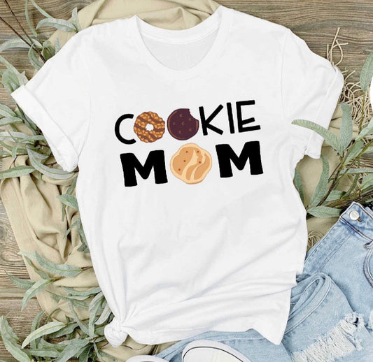 White t-shirt with 'COOKIE MOM' text and cookie graphics on a wooden surface with greenery.