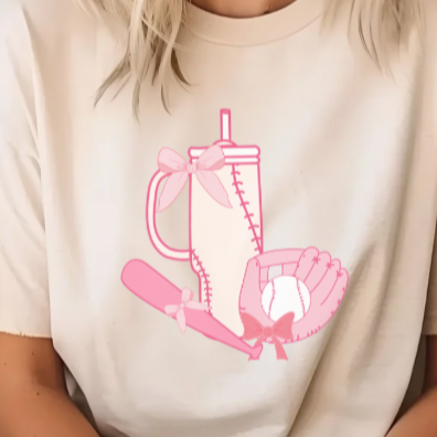 A woman sitting with a beige unisex T-shirt featuring a pink graphic of a girl with a baseball and a cup, and the text 'the cozy season'.