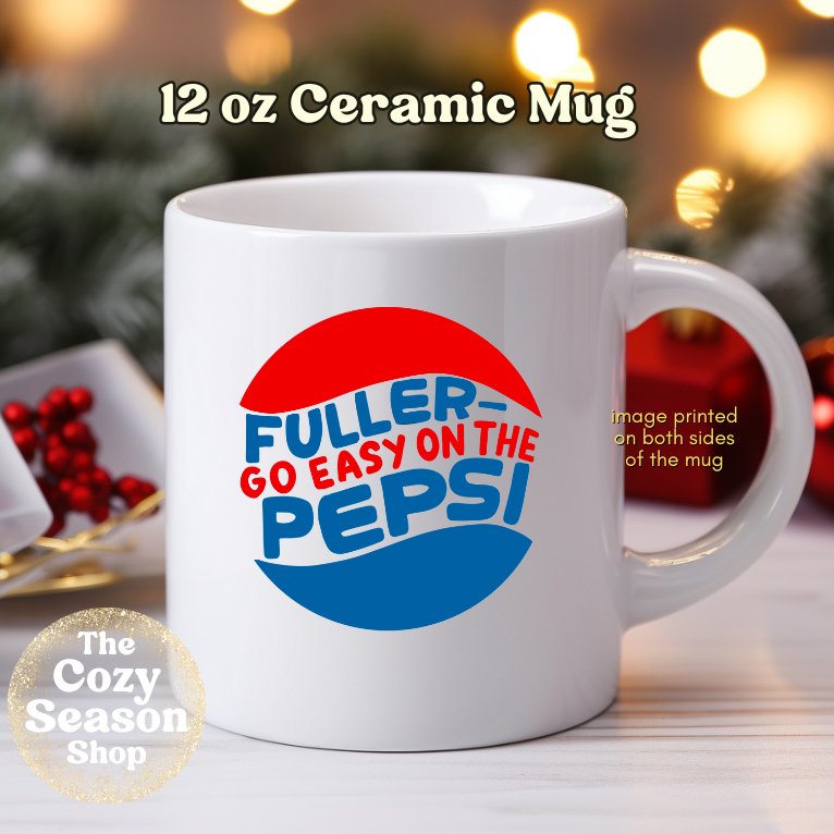 A white 12 oz ceramic mug with a blue and red 'Fuller Go Easy on the Pepsi' design printed on it, displayed on a table with Christmas decorations in the background.