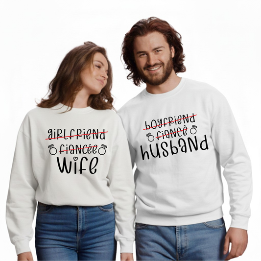 Two people wearing matching white sweatshirts with text (girlfriend, fiancée, WIFE, with girlfriend and fiancée crossed out in red) and (boyfriend, fiance, HUSBAND, with boyfriend and fiancé crossed out in red) , standing side by side on a white background.