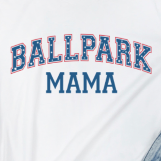 A white unisex crew shirt with 'Ballpark Mama' text and a blue and red graphic on the front, displayed on a wooden surface with a pair of jeans and white sandals.