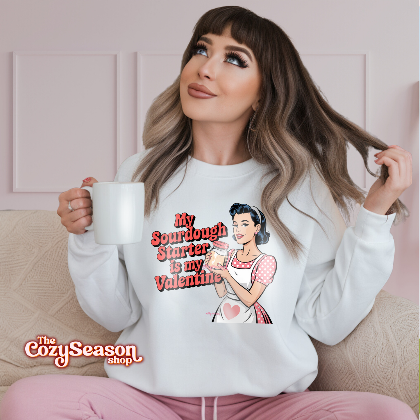 A WOMAN WEARING A WHITE SWEATSHIRT WITH A COMIC ART STYLE DRAWING OF A PIN UP GIRL WEARING A RED AND WHITE POLKADOT DRESS AND A WHITE APRON. THE PIN UP GIRL IS HOLDING A JAR OF SOURDOUGH STARTER. THE RED TEXT: MY SOURDOUGH STARTER IS MY VALENTINE 