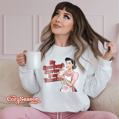A WOMAN WEARING A WHITE SWEATSHIRT WITH A COMIC ART STYLE DRAWING OF A PIN UP GIRL WEARING A RED AND WHITE POLKADOT DRESS AND A WHITE APRON. THE PIN UP GIRL IS HOLDING A JAR OF SOURDOUGH STARTER. THE RED TEXT: MY SOURDOUGH STARTER IS MY VALENTINE 