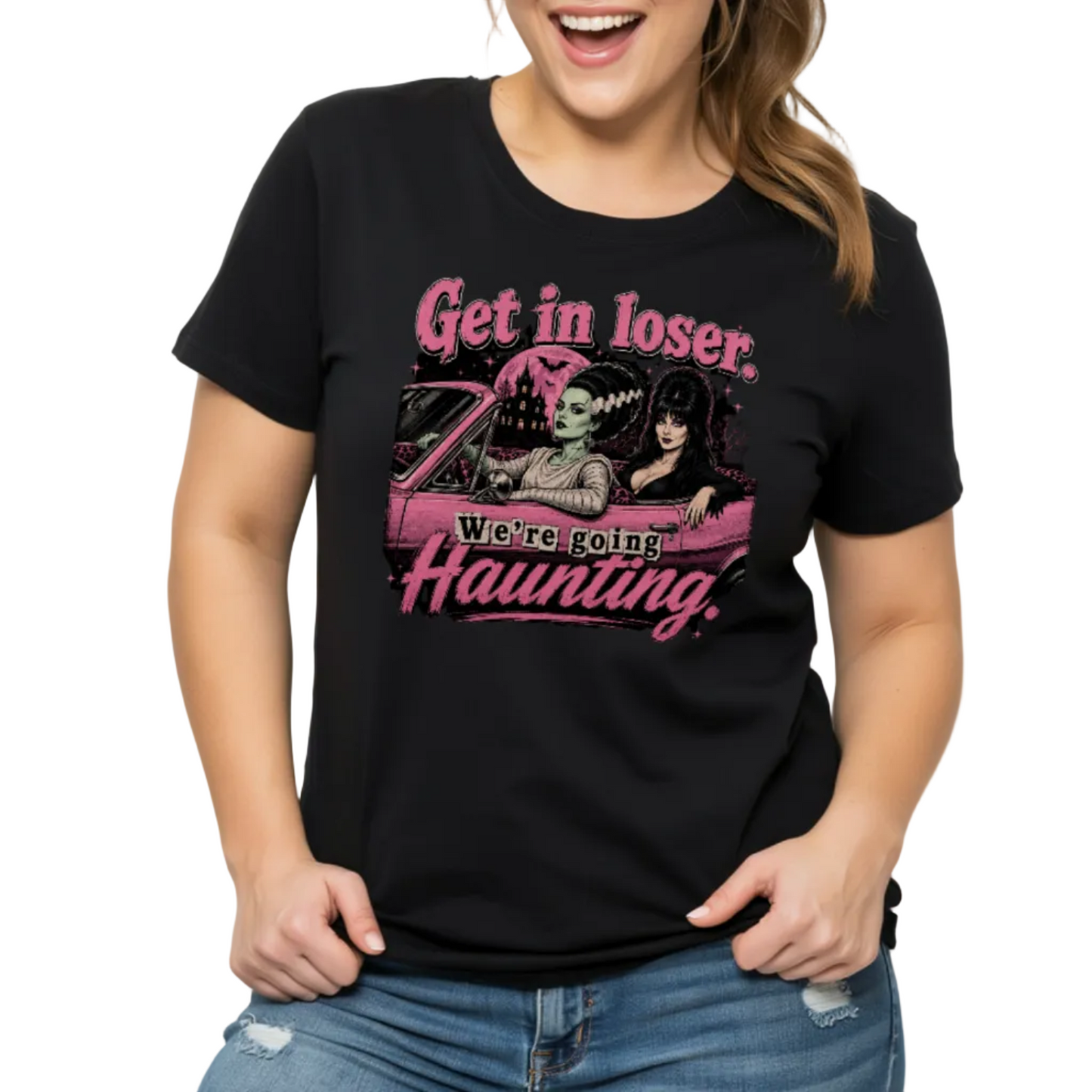 A woman wearing a black graphic t-shirt with a pink and white 'Get in loser. We're going! Haunting!' design, paired with blue jeans, against a plain white background.