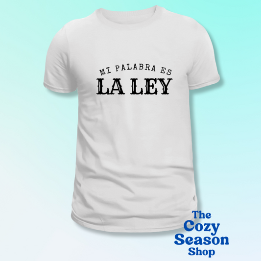 White t-shirt with black text "MI PALABRA ES LA LEY" on a light blue background, featuring 'The Cozy Season Shop' branding.