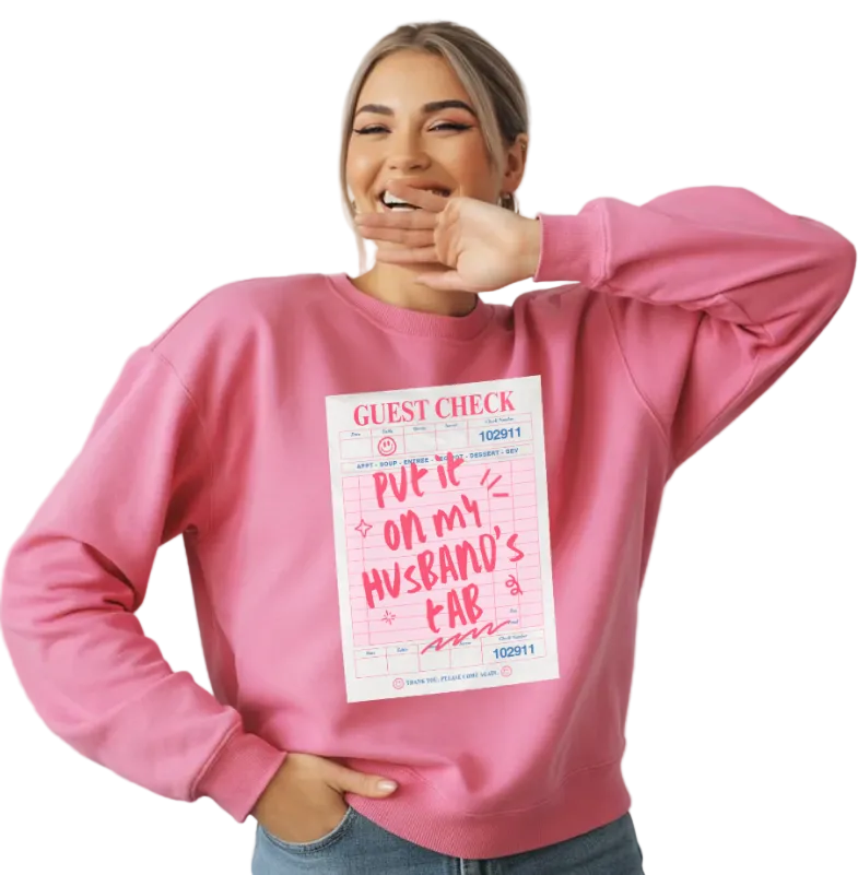 Person wearing a pink sweatshirt with a printed design of a restaurant "Guest Check" with "PUT IT ON MY HUSBAND'S TAB" is handwritten in hot pink across the paper. 