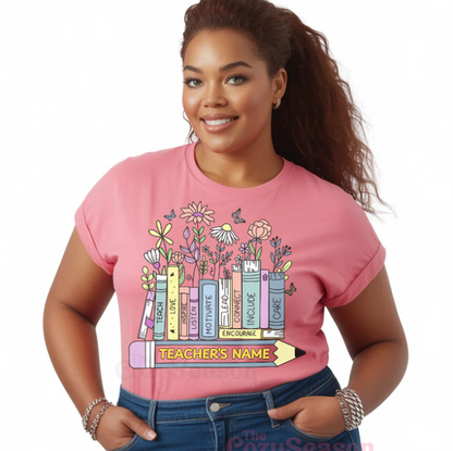 PINK TSHIRT WITH A DESIGN OF BOOKS. EACH BOOK HAS DIFFERENT MOTIVATIONAL WORDS: Teach, love, inspire, listen, motivate, lead, connect, include, encourage, care. Under the books is a large pencil with an empty space for personalization for a teacher's name.  The books have flowers growing out of them on top as a few butterflies flutter about.