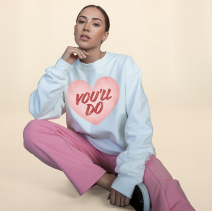 Person wearing a white sweatshirt with a pink heart design and text, sitting on a beige background.