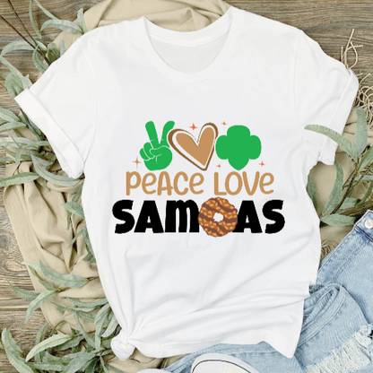 A white unisex adult t-shirt with the text 'PEACE LOVE SAMOAS' and a green leaf logo, displayed on a wooden background with a pair of blue jeans and a pair of white sneakers to the side.