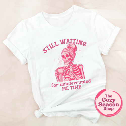 A white short-sleeved t-shirt with a graphic print of a skeleton and the text 'Still waiting for my uninterrupted me time' on the front.