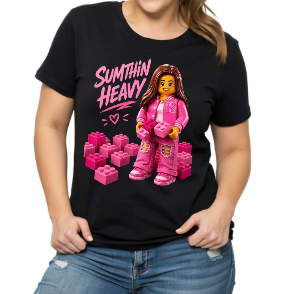 A woman wearing a black short-sleeve graphic T-shirt with a pink LEGO-style minifigure and the words 'SUTMTHIN HEAVY' printed on the front, paired with blue jeans against a plain white background.